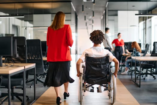 A business leader with her colleague, an African-American businessman who is a disabled person, pass by their colleagues who work in modern offices.