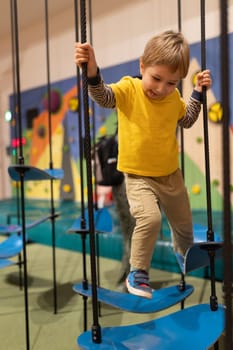 A young boy is hanging from a rope and balancing on a blue platform. The scene is set in a playground with a wall in the background