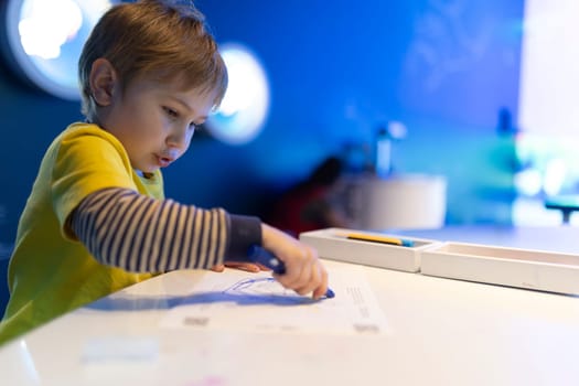 A young boy is drawing on a piece of paper with a blue crayon. He is wearing a yellow shirt and striped long sleeve shirt