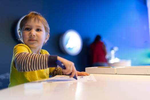 A young boy is sitting at a table with a blue marker in his hand. He is looking at a piece of paper and seems to be drawing or writing something. Concept of creativity and focus
