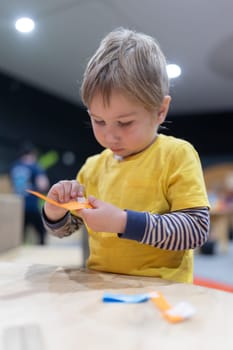 A young boy is sitting at a table, cutting paper with scissors. He is wearing a yellow shirt and has a serious expression on his face. The scene suggests that the boy is focused on his task