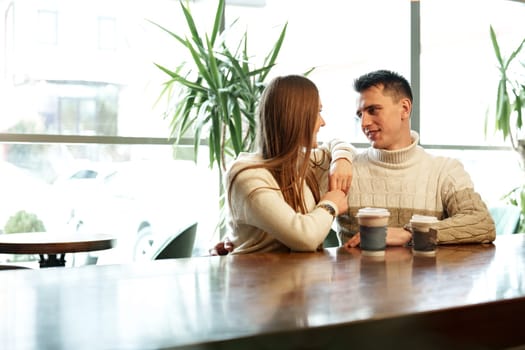 A couple sits close together at a wooden table in a brightly lit cafe, exchanging smiles over steaming cups of perhaps coffee or tea. They appear relaxed and engaged in a cheerful conversation, with natural light filtering through the space creating a warm and inviting atmosphere.