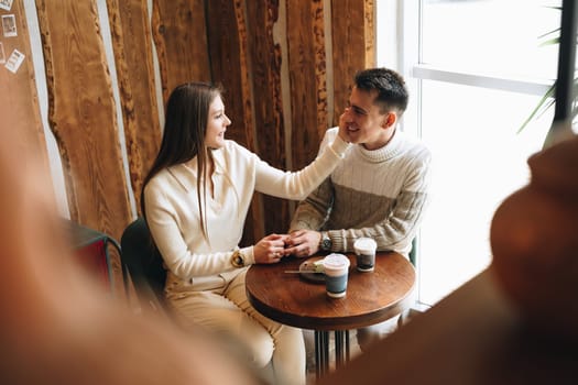 A woman and a man are sitting at a wooden table in a warmly lit cafe, their faces animated in what appears to be a relaxed conversation. The woman holds a paper cup, possibly containing coffee, as they both enjoy a comfortable moment together, surrounded by an ambiance of casual chatter.