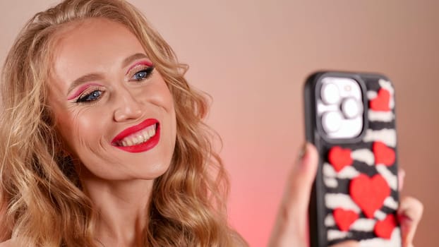 A young woman with red lipstick takes a selfie with long blond hair and makeup. The girl is smiling and looking at the camera close-up