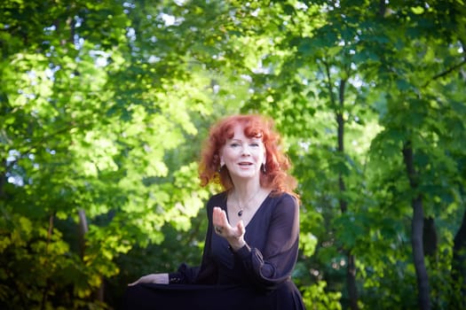 Elegant senior mature Woman dancing in Black Dress in green park. Woman with red hair posing on nature