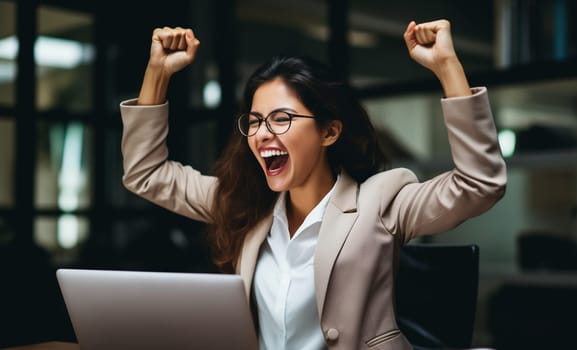 Happy overjoyed laughing businesswoman celebrating success raising his hands up while working on laptop sitting at his desk in the office