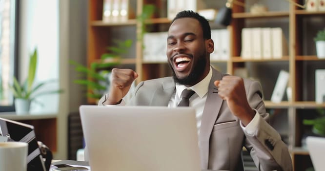 Happy overjoyed laughing african businessman celebrating success raising his hands up while working on laptop sitting at his desk in the office