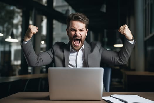 Happy overjoyed laughing businessman celebrating success raising his hands up while working on laptop sitting at his desk in the office