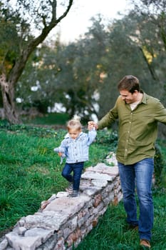 Dad leads a little girl along a stone fence in the park. High quality photo