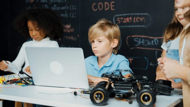 Closeup of boy using laptop programing engineering code and writing program while group of smart diverse student standing surrounded by friend in STEM technology classroom at blackboard. Erudition.