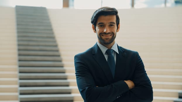 Smart caucasian business man standing with folded arm while looking at camera. Professional handsome project manager pose at camera while smiling with confident with blurred background. Exultant.