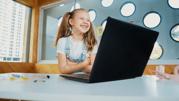 Cute girl using laptop programing software while looking and laughing at camera. Pretty child smiling while crossing arms with funny innocent mood in Stem class. Smart student portrait.Erudition.