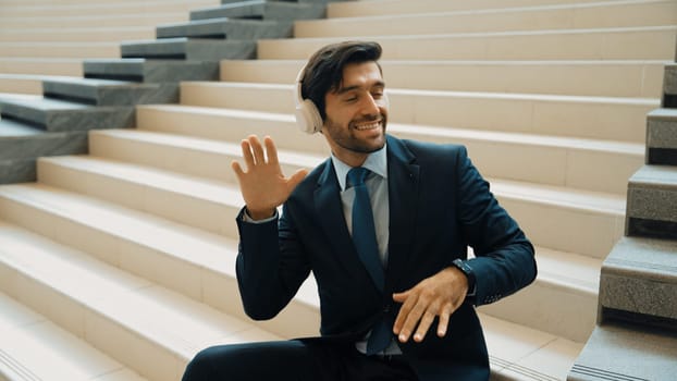 Project manager listening and enjoy music from headphone while sitting at stairs. Professional businessman wearing suit outfit while dancing and moving to music. Happy man listen funny song. Exultant.