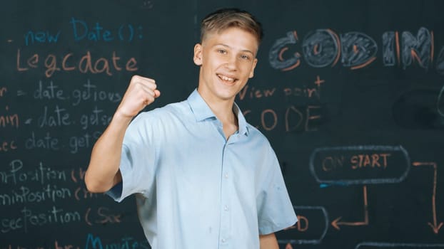 Handsome adult standing at blackboard with engineering code or prompt while attend in STEM class. Student raised hand to celebrate successful project or product while looking at camera. Edification.