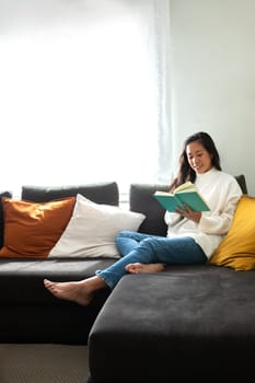 Portrait of happy, smiling Asian woman relaxing at home reading a book sitting on the sofa. Vertical image. Copy space. Lifestyle concept.