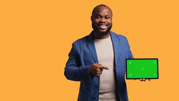 Upbeat african american man holding green screen tablet, doing recommendation. Cheerful BIPOC person pointing towards mockup device, giving positive feedback, studio background, camera B