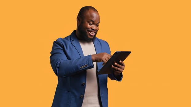 Smiling man scrolling on tablet touchscreen, using internet and credit card to pay for products. African american person happily browsing online shopping websites, studio background, camera B