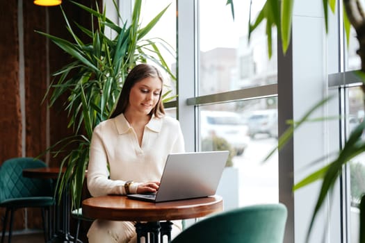 A woman is seated at a table, focused on her laptop computer. She appears to be typing and scrolling through the screen, possibly working or studying.