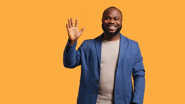 African american man cheerfully doing salutation hand gesture. Portrait of happy BIPOC person raising arm to greet someone, gesturing, isolated over yellow studio background, camera A