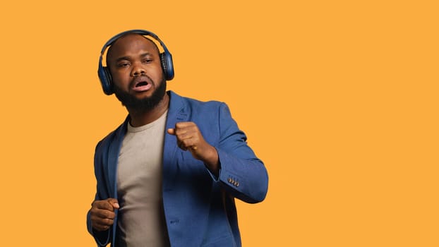 Carefree man listening energic music, raving on rhythm, enjoying free time. Euphoric BIPOC person wearing headphones, doing dance movements, isolated over studio background, camera A