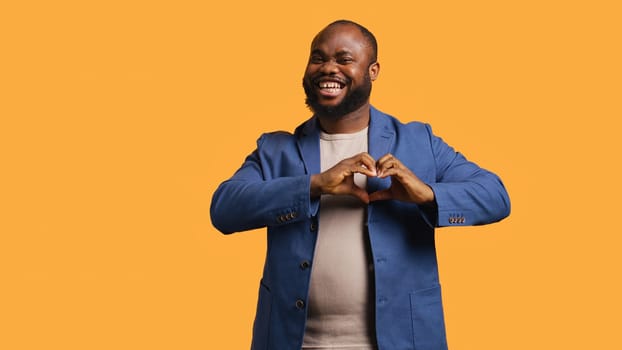 Portrait of jolly friendly african american man doing heart symbol shape gesture with hands. Cheerful nurturing BIPOC person showing love gesturing, isolated over studio background, camera B