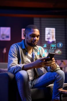 African american man at home using mobile applications on phone and AR technology. BIPOC internet user relaxing on couch, spending time online on smartphone, enjoying leisure time