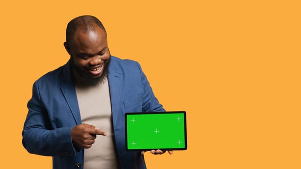 Upbeat african american man holding green screen tablet, doing recommendation. Cheerful BIPOC person pointing towards mockup device, giving positive feedback, studio background, camera A