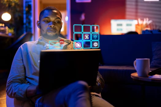 BIPOC teleworker typing on laptop keyboard, doing business tasks at home, accessing digital information concept. Man on couch using AR tech to interact with notebook, app icons on grid visualization