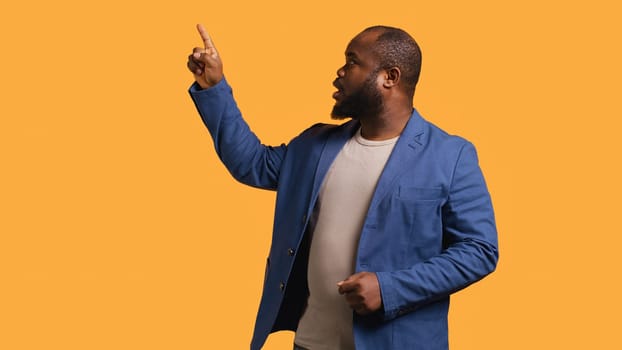 Salesman pointing upwards with finger doing recommendation, talking with audience. BIPOC person looking up, showing copy text, isolated over yellow studio background, camera B