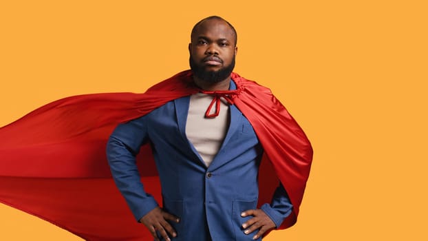 African american superhero flying with red cape, isolated over studio background, flexing muscles. Man wearing cloak posing as hero in costume showing courage and strength, camera B