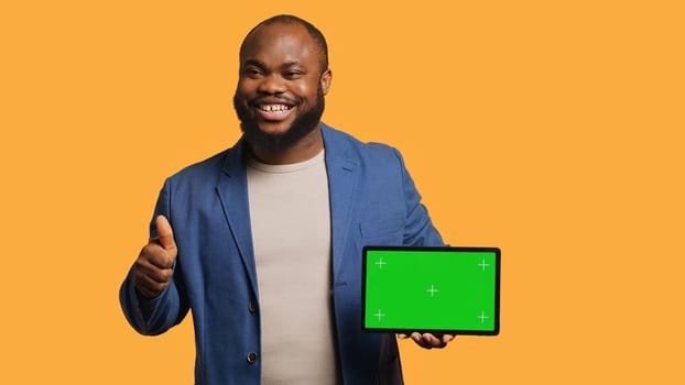 Jolly man holding isolated screen tablet, doing recommendation. Cheerful BIPOC person pointing towards chroma key device, doing thumbs up sign, studio background, camera A