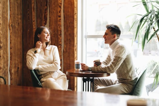 A woman and a man are sitting at a wooden table in a warmly lit cafe, their faces animated in what appears to be a relaxed conversation. The woman holds a paper cup, possibly containing coffee, as they both enjoy a comfortable moment together, surrounded by an ambiance of casual chatter.