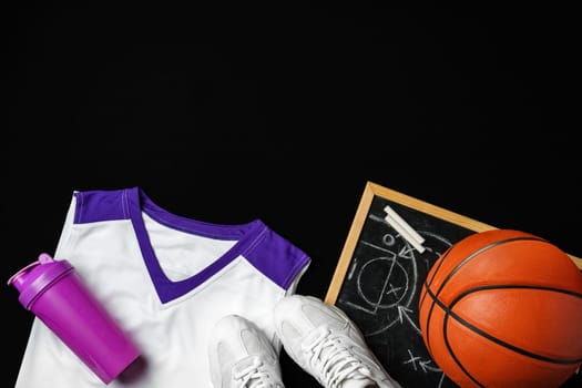 A collection of basketball essentials is neatly arranged against a stark black backdrop. A pair of white sneakers with purple accents lies next to a vibrant orange basketball. On the right, a clipboard with a basketball court diagram and a strategy sketched in white chalk can be seen, while a white and purple basketball jersey drapes gracefully at the edge. Completing the set, a purple sports water bottle caps off the ensemble, suggesting preparation for an upcoming game or practice session.