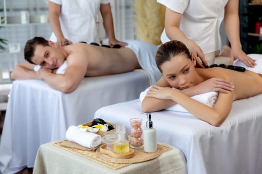 Hot stone massage at spa salon in luxury resort with day light serenity ambient, blissful couple customer enjoying spa basalt stone massage glide over body with soothing warmth. Quiescent