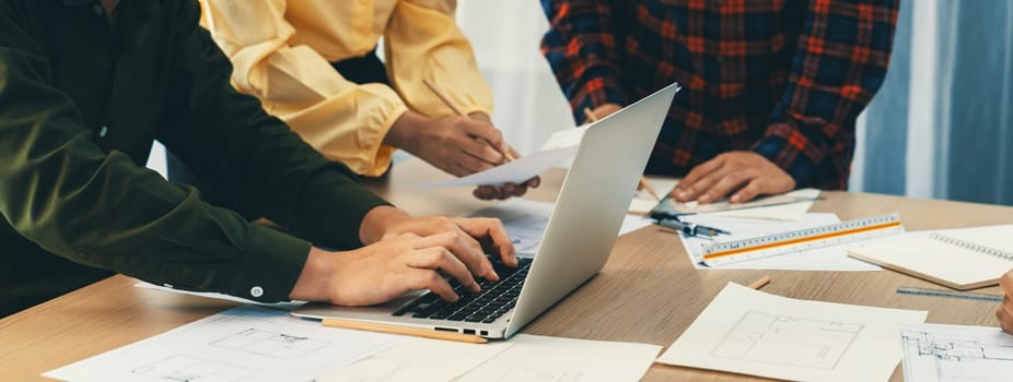 Expert engineer team discussion about construction design while male engineer using laptop analysis data on table with blueprint and architectural equipment scatter around. Closeup. Delineation.