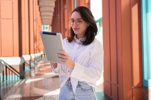 Businesswoman in glasses using digital tablet, standing outdoor. Female freelancer holding tablet while walk outside