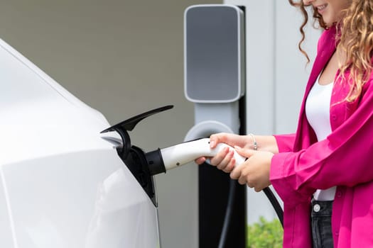 Modern eco-friendly woman recharging electric vehicle from home EV charging station. EV car technology utilized for home resident to future environmental sustainability. Synchronos