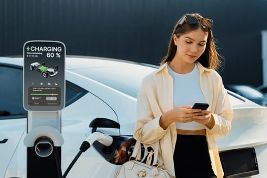 Young woman holding shopping bag and use smartphone to pay for electricity for recharging EV car battery from charging station at city mall parking lot. Modern woman go shopping by eco car. Expedient