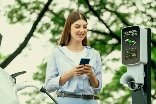 Young woman using smartphone online banking application to pay for electric car battery charging from EV charging station during vacation holiday road trip at national park or summer forest. Exalt