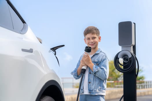 Playful little boy pointing EV charger at camera, recharging eco-friendly electric car from EV charging station. EV car travel by the seashore using clean and sustainable energy.Perpetual