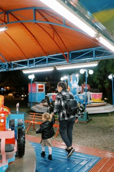 Mom and little girl walk holding hands past colorful carousels. Back view. High quality photo