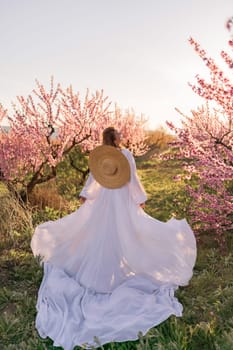 Woman blooming peach orchard. Against the backdrop of a picturesque peach orchard, a woman in a long white dress and hat enjoys a peaceful walk in the park, surrounded by the beauty of nature