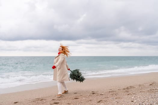 Redhead woman Christmas tree sea. Christmas portrait of a happy redhead woman walking along the beach and holding a Christmas tree in her hands. Dressed in a light coat, white suit and red mittens