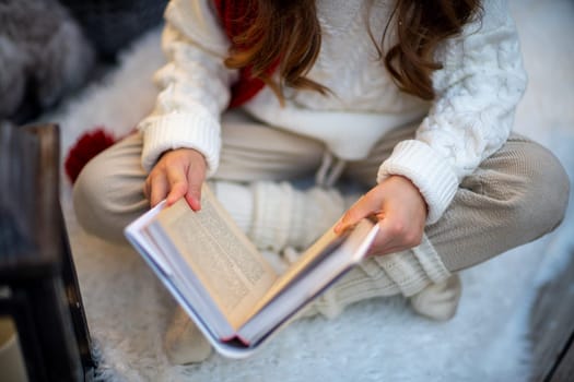 A girl with a book is sitting on the floor on a furry white carpet. Dressed in a white sweater and light trousers with a red scarf