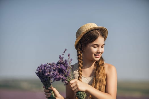 girl is holding a bunch of lavender purple flowers in her hands and wearing a straw hat. She is smiling and she is enjoying herself. The scene is set in a field of lavender, which adds to the peaceful
