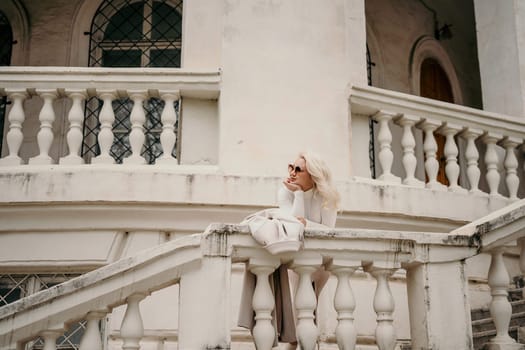 A woman is sitting on a white railing, looking out at the street. She is wearing sunglasses and a white shirt. The scene is set in front of a building with white pillars