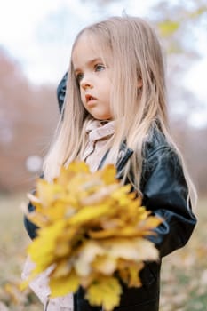 Little girl with a bouquet of yellow leaves stands in the park and scratches her head. High quality photo
