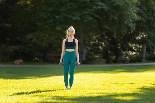 A woman is standing in a park, holding two dumbbells. She is wearing a green top and green pants. The park is filled with trees and grass, and there is a bench nearby