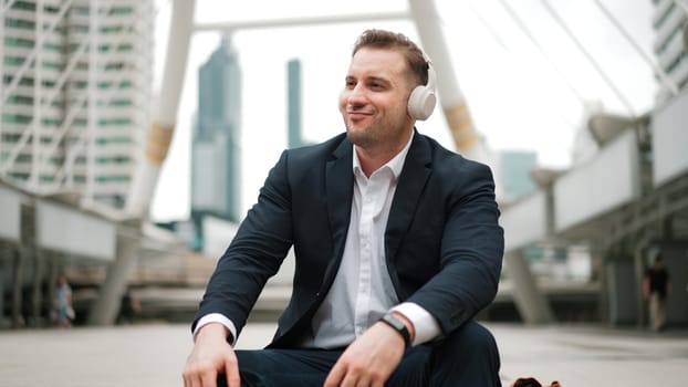 Smart business man wear headphone while sitting at stair in urban city and listen to relaxing music. Happy male leader using headset and moving to song in lively mood surrounded by skyscraper. Urbane.