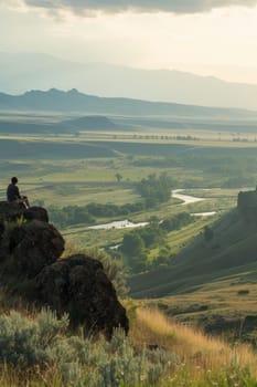 Individual sitting in solitude on a hill, overlooking a tranquil valley with a river snaking through it at dusk